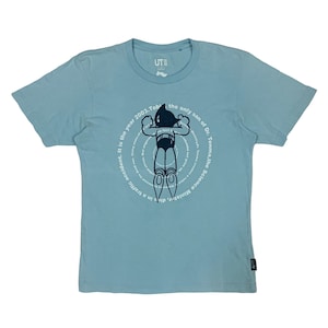 May include: Light blue t-shirt featuring a graphic of a robot with the text "UT" and a circular design with text. The text includes "It is the year 2003. Tobi the only son of Dr. Tenma, the Science Minister, dies in a traffic accident."