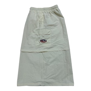 May include: A cream-colored midi skirt featuring an elastic waistband, a horizontal seam across the middle, and a single cargo pocket on the right side. The pocket is adorned with a small, oval-shaped Fila logo patch in red, white, and blue.