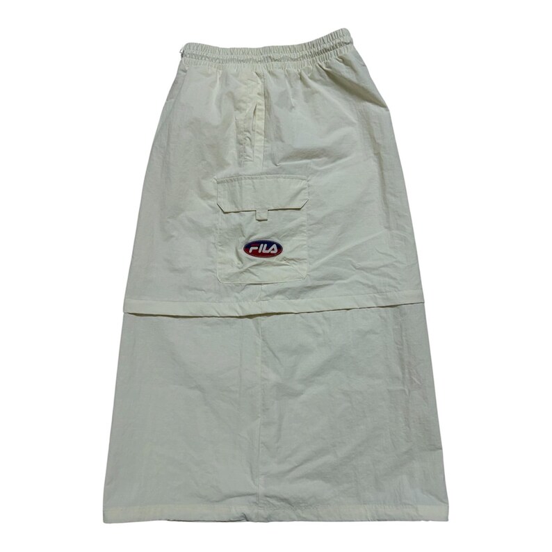 May include: A cream-colored midi skirt featuring an elastic waistband, a horizontal seam across the middle, and a single cargo pocket on the right side. The pocket is adorned with a small, oval-shaped Fila logo patch in red, white, and blue.