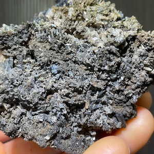 May include: A close-up of a mineral specimen featuring a matrix of dark gray and black crystals. The surface is textured with clusters of smaller, clear and white crystals. The overall appearance is rough and natural.