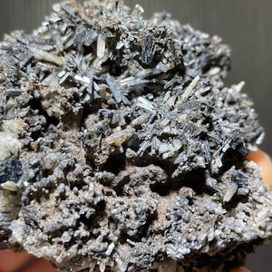 May include: Close-up of a mineral specimen featuring a cluster of dark, needle-like crystals. The crystals are a dark gray color, contrasting with the lighter, earthy-toned matrix. The overall texture is rough and uneven, showcasing the natural formation of the mineral.