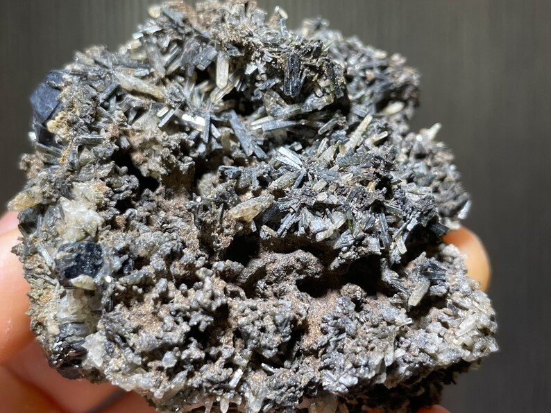 May include: Close-up of a mineral specimen featuring a cluster of dark, needle-like crystals. The crystals are a dark gray color, contrasting with the lighter, earthy-toned matrix. The overall texture is rough and uneven, showcasing the natural formation of the mineral.