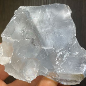 K&ouml;nnte beinhalten: Ein durchscheinendes, hellblaues Mineralexemplar mit einer rauen, unregelm&auml;&szlig;igen Form. Der Stein hat eine kristalline Struktur mit sichtbaren inneren Br&uuml;chen und einem leicht irisierenden Glanz. Das Gesamtbild ist k&uuml;hl und glatt.