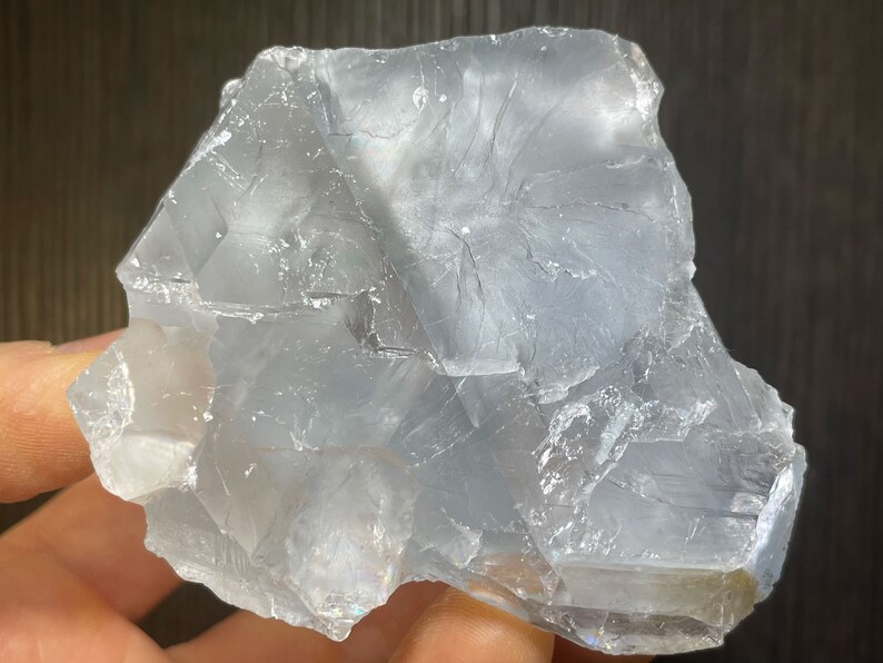 K&ouml;nnte beinhalten: Ein durchscheinendes, hellblaues Mineralexemplar mit einer rauen, unregelm&auml;&szlig;igen Form. Der Stein hat eine kristalline Struktur mit sichtbaren inneren Br&uuml;chen und einem leicht irisierenden Glanz. Das Gesamtbild ist k&uuml;hl und glatt.