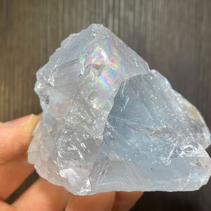 K&ouml;nnte beinhalten: Eine durchscheinende, hellblaue Kristallformation mit rauer, strukturierter Oberfl&auml;che. Der Kristall zeigt schillernde Regenbogenfarben, wo sich das Licht durch seine Facetten bricht. Die Gesamtform ist eckig und unregelm&auml;&szlig;ig.