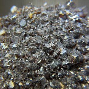 Peut inclure: Gros plan sur un amas de minéraux cristallins sombres. Les cristaux sont principalement gris foncé et noirs, avec une surface légèrement réfléchissante. La texture générale est rugueuse et dentelée, avec de nombreux petits pointes vives. La formation minérale semble dense et compacte.