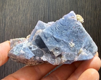 May include: A hand holds a cluster of blue fluorite crystals. The crystals have a layered, geometric structure with a soft, matte finish. Small, light-colored mineral deposits are visible on the surface. The background is a dark wood grain.
