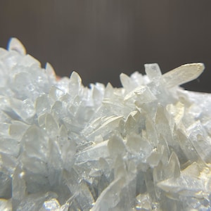 May include: Close-up of a cluster of translucent, crystalline formations. The crystals are a pale, icy white, with sharp, pointed edges. The overall texture is rough and jagged, with a dark background.