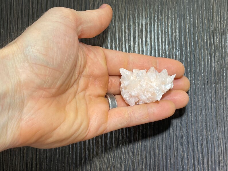 May include: A cluster of light pink selenite crystals, held in a hand. The crystals have a textured, layered appearance, with multiple points and facets. The selenite is a soft, translucent color.