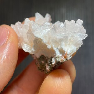 May include: A cluster of white, crystalline formations, resembling a collection of delicate, pointed structures. The mineral specimen is set against a backdrop of a darker, earthy base, creating a contrast in color and texture. The crystals are held by a hand.