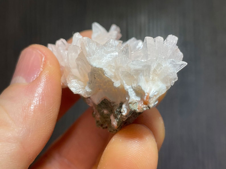 May include: A cluster of white, crystalline formations, resembling a collection of delicate, pointed structures. The mineral specimen is set against a backdrop of a darker, earthy base, creating a contrast in color and texture. The crystals are held by a hand.