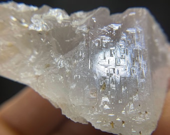 May include: Close-up of a crystalline mineral specimen. The stone is a pale, translucent color with a textured surface. The crystal structure is visible, with geometric patterns and reflective facets.