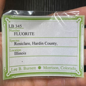 K&ouml;nnte beinhalten: Eine transparente Plastikt&uuml;te mit einem wei&szlig;-gr&uuml;nen Etikett mit dem Text "FLUORITE". Das Etikett enth&auml;lt den Ort "Illinois" und den Namen "Lee B. Burnett, Morrison, Colorado."
