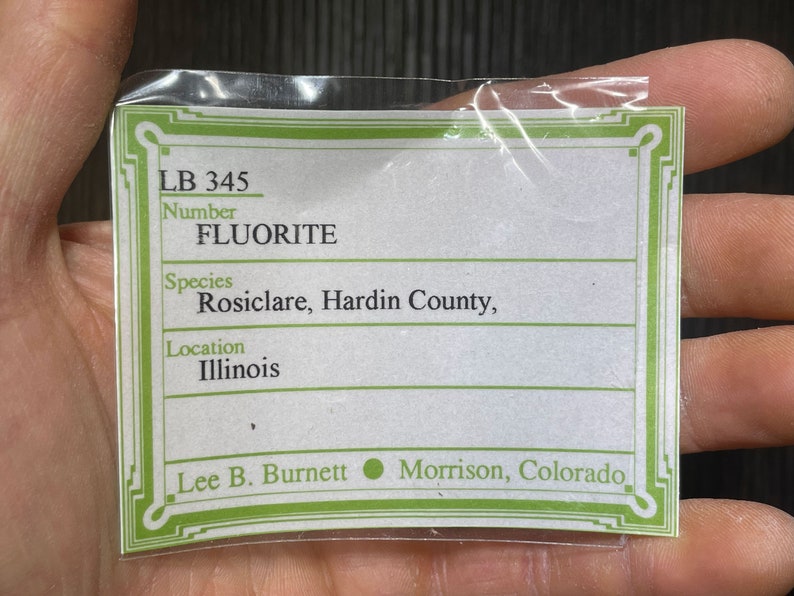 K&ouml;nnte beinhalten: Eine transparente Plastikt&uuml;te mit einem wei&szlig;-gr&uuml;nen Etikett mit dem Text "FLUORITE". Das Etikett enth&auml;lt den Ort "Illinois" und den Namen "Lee B. Burnett, Morrison, Colorado."