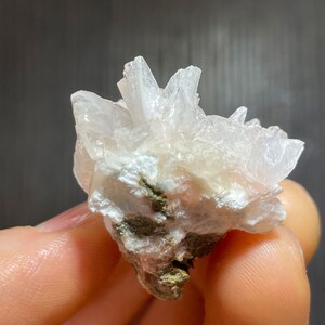 May include: A cluster of white crystal formations with a slightly pinkish hue. The crystals have a sharp, pointed structure and are attached to a base with green and brown mineral deposits. The crystals are held by a person.
