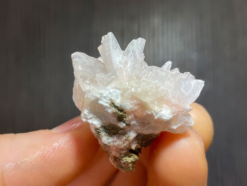 May include: A cluster of white crystal formations with a slightly pinkish hue. The crystals have a sharp, pointed structure and are attached to a base with green and brown mineral deposits. The crystals are held by a person.