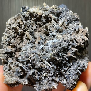 May include: A close-up of a mineral specimen featuring a cluster of dark, needle-like crystals. The crystals are primarily black and gray, with a rough, textured matrix. The overall appearance is rugged and natural, showcasing the raw beauty of the mineral.