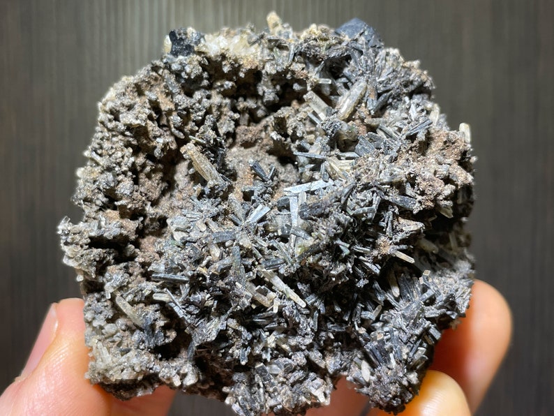 May include: A close-up of a mineral specimen featuring a cluster of dark, needle-like crystals. The crystals are primarily black and gray, with a rough, textured matrix. The overall appearance is rugged and natural, showcasing the raw beauty of the mineral.