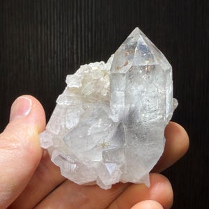 May include: A clear quartz crystal cluster with a large, pointed central crystal and smaller, attached crystals. The quartz has a translucent appearance with subtle internal patterns and inclusions. The crystals are held in a hand.