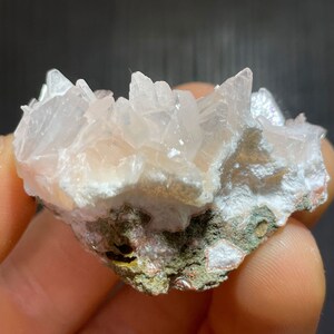 May include: A cluster of pale pink quartz crystals, with sharp, pointed terminations, sits atop a matrix of white and green minerals. The crystals have a translucent quality, catching the light.