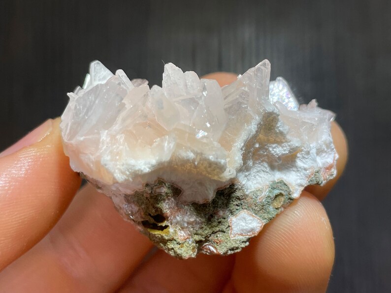 May include: A cluster of pale pink quartz crystals, with sharp, pointed terminations, sits atop a matrix of white and green minerals. The crystals have a translucent quality, catching the light.