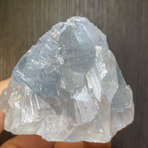 K&ouml;nnte beinhalten: Ein hellblauer Celestit-Kristallcluster. Der Kristall hat eine geschichtete, geometrische Struktur mit sichtbaren Streifen. Die Gesamtform ist unregelm&auml;&szlig;ig, mit einer rauen, strukturierten Oberfl&auml;che. Die Farbe ist ein blasses, durchscheinendes Blau.