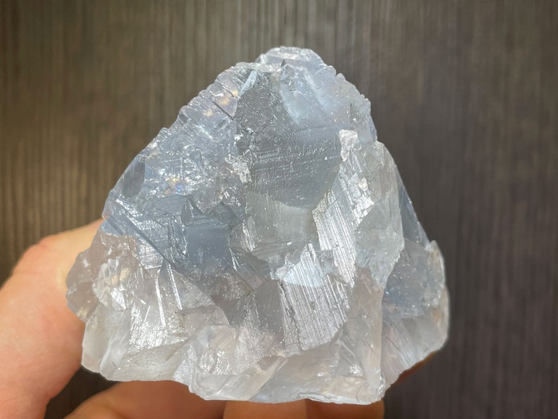 K&ouml;nnte beinhalten: Ein hellblauer Celestit-Kristallcluster. Der Kristall hat eine geschichtete, geometrische Struktur mit sichtbaren Streifen. Die Gesamtform ist unregelm&auml;&szlig;ig, mit einer rauen, strukturierten Oberfl&auml;che. Die Farbe ist ein blasses, durchscheinendes Blau.