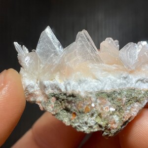 May include: A close-up of a cluster of translucent, crystalline formations in shades of white and peach. The crystals are arranged on a base of white and green rock, creating a natural, textured appearance. The mineral specimen is held by a person.