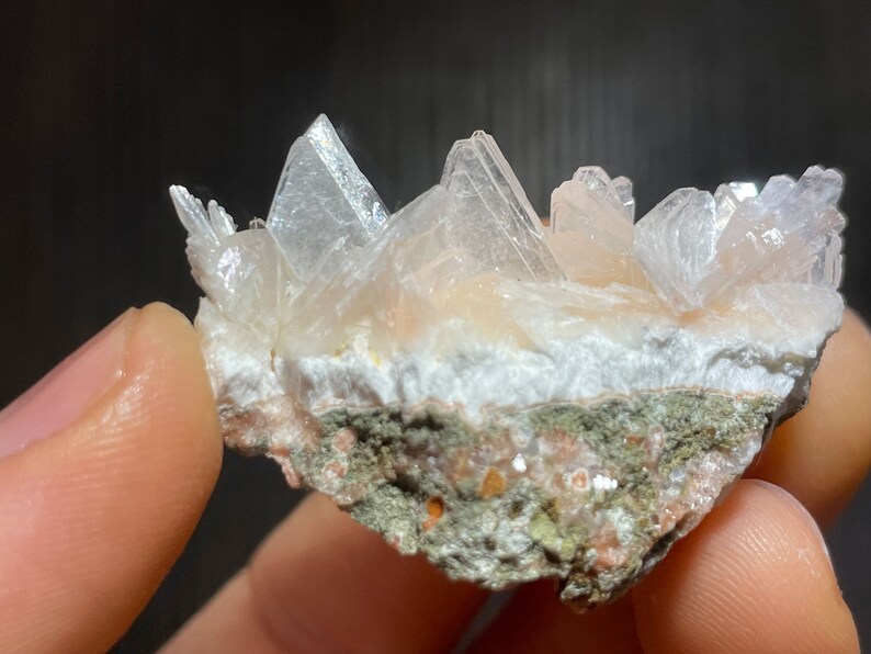 May include: A close-up of a cluster of translucent, crystalline formations in shades of white and peach. The crystals are arranged on a base of white and green rock, creating a natural, textured appearance. The mineral specimen is held by a person.
