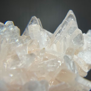 May include: Close-up of a cluster of translucent, pale pink crystals. The crystals have sharp, pointed tips and a layered structure, creating a textured and intricate appearance. The background is a dark gray.