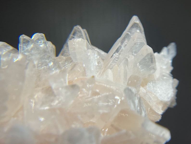 May include: Close-up of a cluster of translucent, pale pink crystals. The crystals have sharp, pointed tips and a layered structure, creating a textured and intricate appearance. The background is a dark gray.