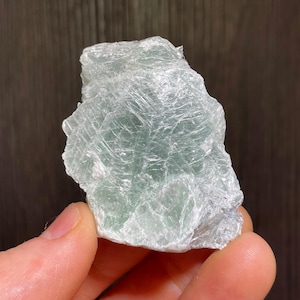May include: A rough fluorite crystal, pale green and white in colour. The mineral has a textured surface and is held in a hand. The crystal appears translucent, with visible internal structures. A natural, uncut gemstone.