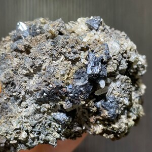 May include: A close-up of a mineral specimen featuring a matrix of gray and brown rock. Embedded within the matrix are several shiny, metallic-looking, dark gray crystals. Some clear, crystalline structures are also visible.
