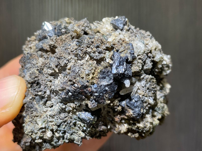 May include: A close-up of a mineral specimen featuring a matrix of gray and brown rock. Embedded within the matrix are several shiny, metallic-looking, dark gray crystals. Some clear, crystalline structures are also visible.