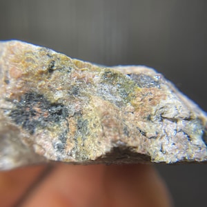 May include: Close-up of a rough-hewn rock fragment. The stone displays a mix of colors, including tan, pink, black, and green. The texture appears granular and uneven, with visible mineral inclusions. The overall appearance suggests a natural, unpolished specimen.