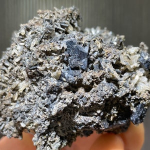 May include: A close-up of a mineral specimen featuring a cluster of dark gray, crystalline structures. The specimen has a rough, textured surface with small, light-colored crystal formations throughout. The overall color palette is a mix of gray, black, and off-white.