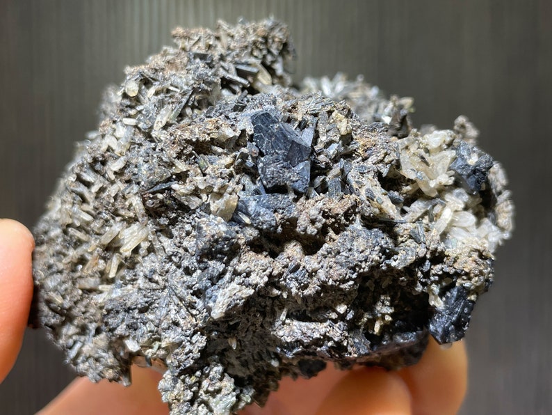 May include: A close-up of a mineral specimen featuring a cluster of dark gray, crystalline structures. The specimen has a rough, textured surface with small, light-colored crystal formations throughout. The overall color palette is a mix of gray, black, and off-white.