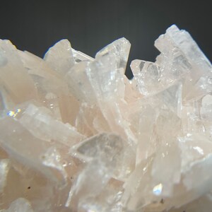 May include: Close-up of a cluster of pale pink crystal formations. The crystals are sharp and angular, with a translucent quality. The background is a dark gray, providing contrast to the light-colored crystals.