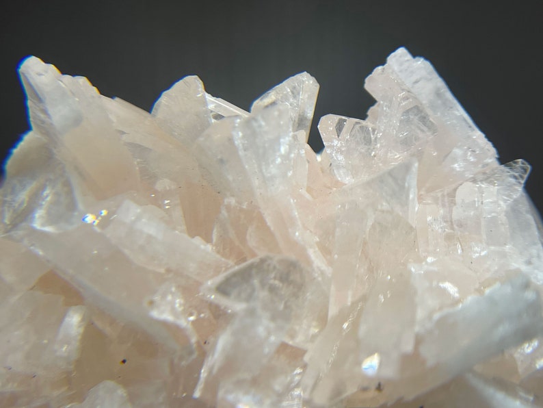 May include: Close-up of a cluster of pale pink crystal formations. The crystals are sharp and angular, with a translucent quality. The background is a dark gray, providing contrast to the light-colored crystals.