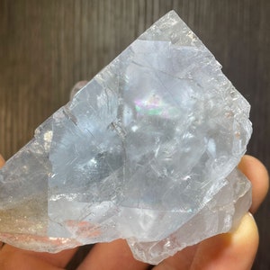 K&ouml;nnte beinhalten: Eine durchscheinende, hellblaue Kristallformation mit irisierenden Bereichen. Der Kristall hat eine raue, unebene Oberfl&auml;che und eine geometrische Form. Der Kristall wird in einer Hand gehalten, mit einem dunkelbraunen Hintergrund.
