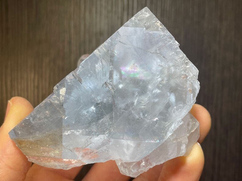 K&ouml;nnte beinhalten: Eine durchscheinende, hellblaue Kristallformation mit irisierenden Bereichen. Der Kristall hat eine raue, unebene Oberfl&auml;che und eine geometrische Form. Der Kristall wird in einer Hand gehalten, mit einem dunkelbraunen Hintergrund.