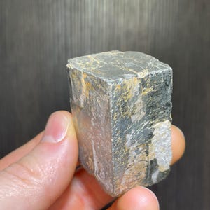 May include: A silver-colored, cube-shaped mineral specimen with a textured surface. The mineral has a metallic sheen and displays areas of gold and black coloration. The specimen is held in a hand, showcasing its size and form.