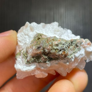 May include: A close-up of a mineral specimen featuring a cluster of white, crystalline formations. The specimen has a central band of green and reddish-brown hues, with a rough, textured appearance. The background is dark.