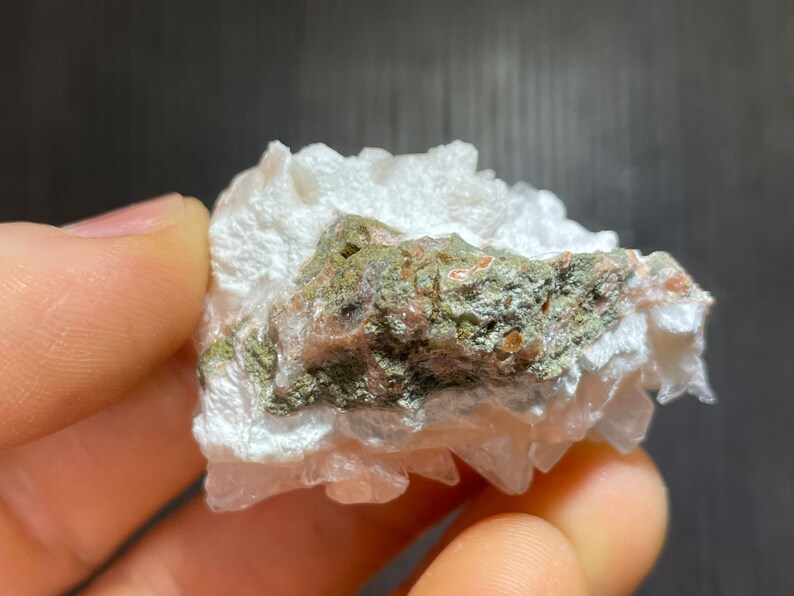 May include: A close-up of a mineral specimen featuring a cluster of white, crystalline formations. The specimen has a central band of green and reddish-brown hues, with a rough, textured appearance. The background is dark.