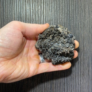 May include: A dark gray mineral specimen with needle-like crystal formations and a rough, textured surface. The mineral is held in a hand, showcasing its natural, unpolished appearance. The color palette is dominated by shades of gray and black.