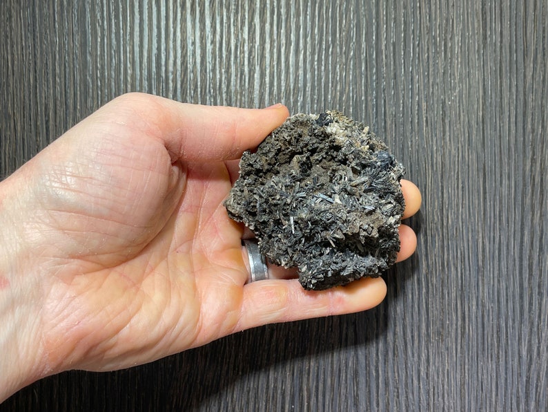 May include: A dark gray mineral specimen with needle-like crystal formations and a rough, textured surface. The mineral is held in a hand, showcasing its natural, unpolished appearance. The color palette is dominated by shades of gray and black.