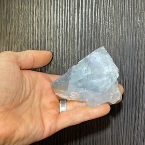 K&ouml;nnte beinhalten: Ein hellblaues, rohes Kristall in einer Hand gehalten. Der Kristall hat eine raue, strukturierte Oberfl&auml;che mit sichtbaren Br&uuml;chen. Der Stein ist hellblau mit einigen durchscheinenden Bereichen. Der Hintergrund ist eine dunkle Holzmaserung.