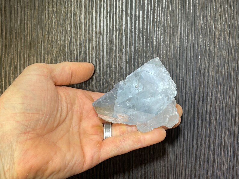 K&ouml;nnte beinhalten: Ein hellblaues, rohes Kristall in einer Hand gehalten. Der Kristall hat eine raue, strukturierte Oberfl&auml;che mit sichtbaren Br&uuml;chen. Der Stein ist hellblau mit einigen durchscheinenden Bereichen. Der Hintergrund ist eine dunkle Holzmaserung.