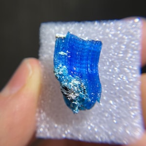 May include: A vibrant blue mineral specimen with a crystalline structure. The mineral has a deep, saturated blue color with visible striations. Small patches of white and dark gray material are present on the surface. The mineral is displayed on a white, textured surface.