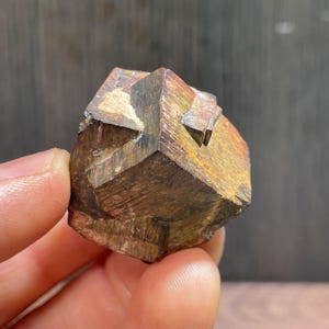 May include: A pyrite cube, a mineral with a metallic sheen. The cube has a geometric form with multiple facets, showing a mix of golden and bronze shades. The mineral is held in a hand, against a dark backdrop.